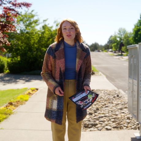 Woman in a plaid coat holding flyers stands on a sunny suburban sidewalk near mailboxes.